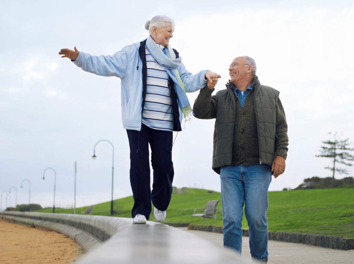 Ett äldre par som går på en strandpromenad. Kvinnan balanserar och mannen håller hennes hand.